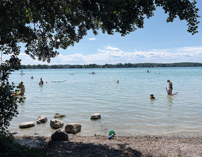 karibik_feeling_am_woerthsee Blick unter Bäumen an einem Kiesstrand auf einen See, in dem gebadet wird