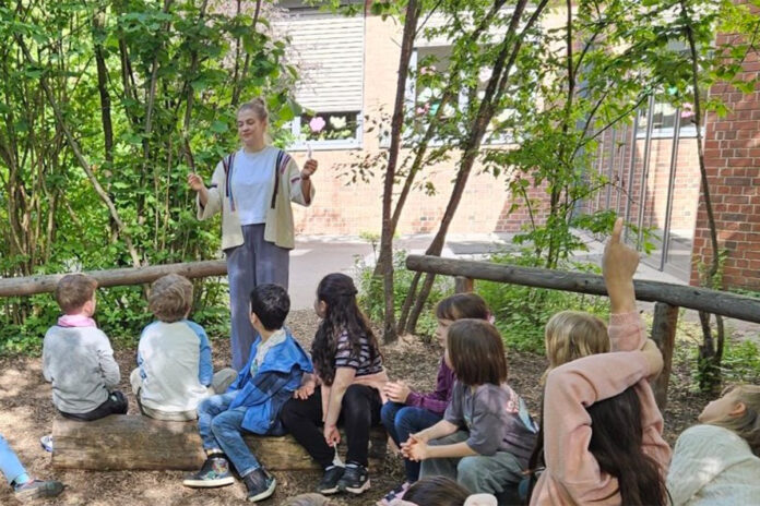 Akasha_Akademy Kinder sitzen auf einen Baumstamm und hören einer stehenden Frau zu, die etwas erklärt